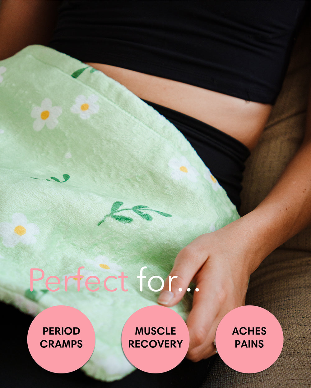 Green heating pad with floral patterns on a person's lap, labeled for period cramps, muscle recovery, and aches/pains.