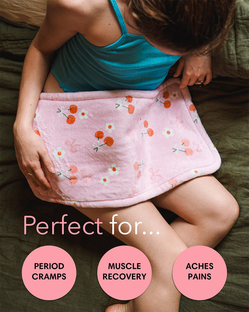 Perfect for period cramps, muscle recovery, and aches. Woman laying on bed using cherry pink cute heating pad for period cramps, pain, and menstrual relief. 
