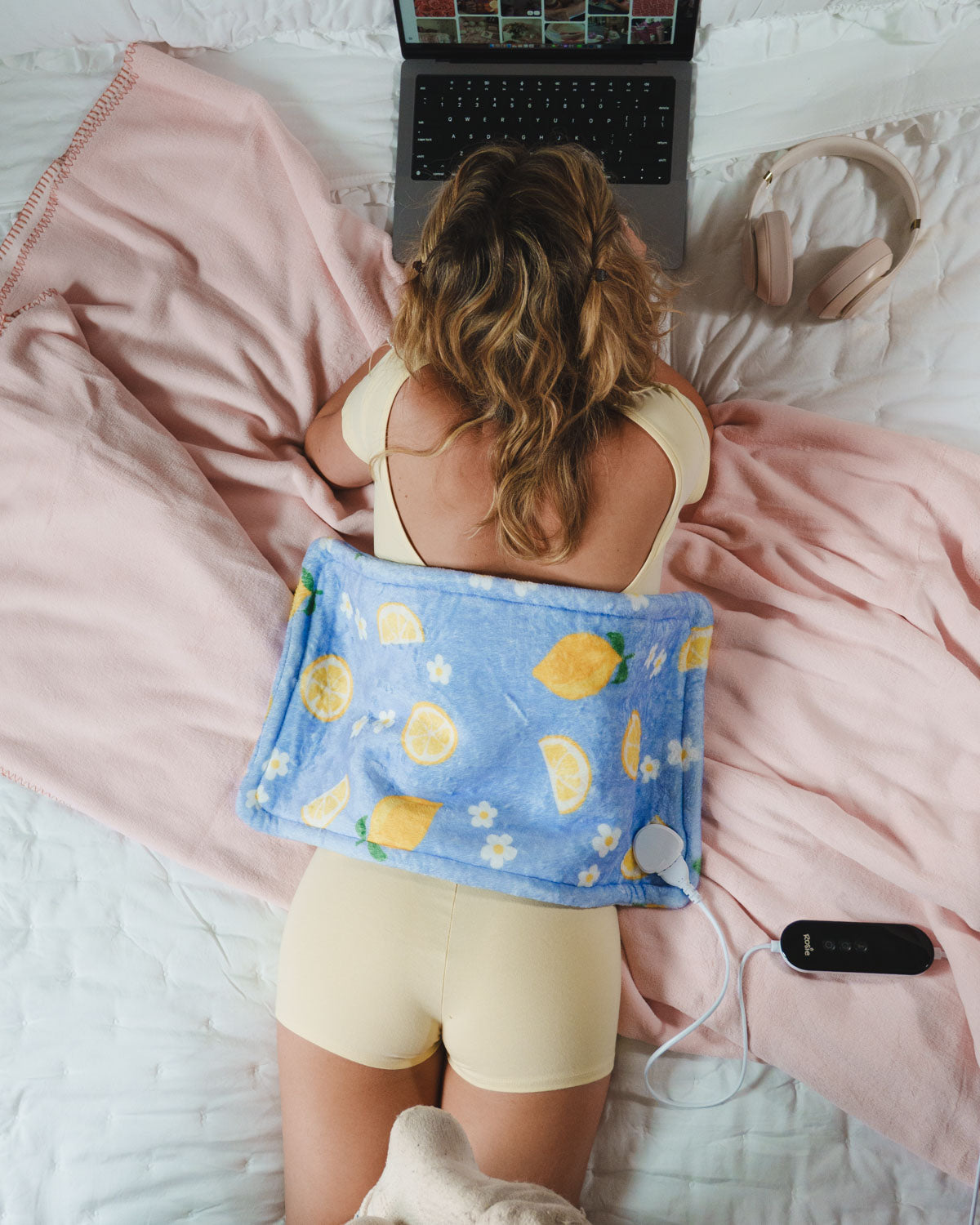 Woman lying on a bed with a blue heating pad featuring lemon patterns, using a laptop.